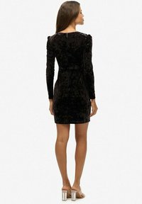 Schwarzes Samtkleid mit langen Ärmeln, taillierter Silhouette, strukturierten Oberfläche und subtiler Glanz. Reißverschluss auf der Rückseite und geraffte Schulterakzente.