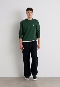 Grønn genser, sorte bukser, hvit skjorte under. Enkelt design, avslappet passform. Genseren har et lite logo; modellen har på seg grå joggesko.