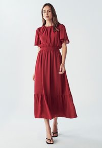 Crvena maksi haljina s kratkim puf rukavima, smotanom strukom i višeslojnim rubom. Proljetno tkanje, glatka tekstura i elegantna silueta.