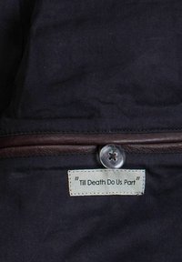 Tume kangas lähivaates, millel on beež etikett tekstiga "Till Death Do Us Part", kinnitatud ümmarguse hõbedase nupu ja nahktriibuga.