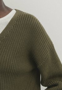 Persona che indossa un cardigan chunky di un verde oliva sopra una camicia bianca con texture, sfondo grigio chiaro semplice.