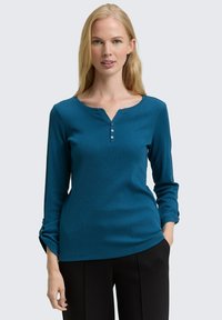 Blonde vrouw in een teal lange mouwen henley shirt met drie knopen, zwarte broek, staande met één hand in de zak.