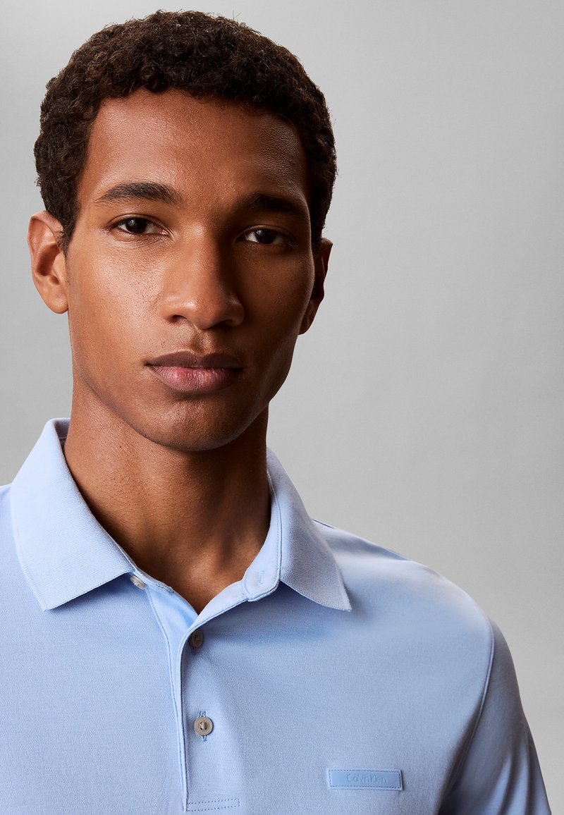 Jonge man met kort krullend haar, gekleed in een lichtblauw Calvin Klein polohemd tegen een effen grijze achtergrond.