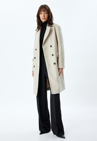 Beige dobbelbrystet frakke med store knapper, brede klapper og en skræddertilpasset pasform. Blødt stof og sidelommer; båret over en sort turtleneck.