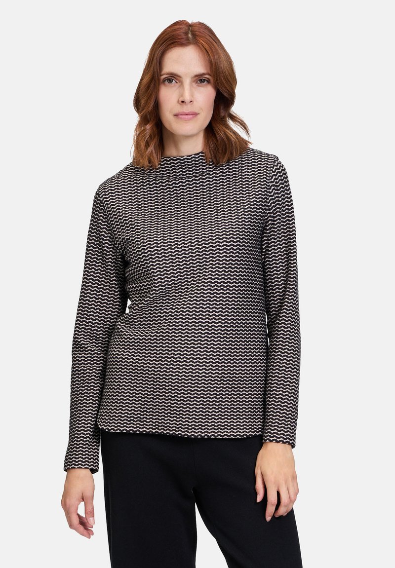 Sort og hvid langærmet top med et struktureret zigzag-mønster, høj halsudskæring og en afslappet pasform. Sammen med mørke bukser.