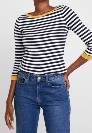 Personne portant une chemise à manches longues rayée noir et blanc avec des poignets et un col jaunes, associée à un jean en denim bleu.