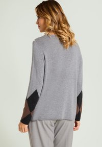 Suéter gris de manga larga con patrones geométricos en negro y marrón en las mangas. Textura suave, ajuste relajado, escote redondo.