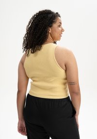 Gelbes, geripptes Tanktop mit taillierter Passform, kombiniert mit schwarzen, locker sitzenden Hosen. Sichtbares Tattoo auf dem linken Arm.