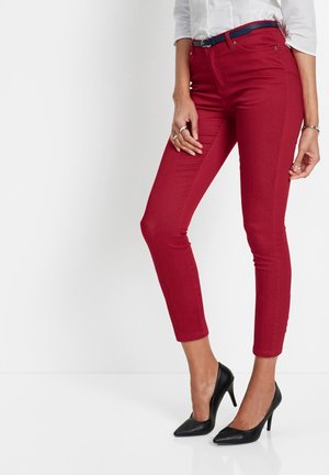 Femme portant un pantalon rouge ajusté et court avec une ceinture bleue, un chemisier blanc, des talons hauts noirs et des bijoux argentés aux mains et au poignet.