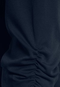 Detailní záběr na tmavě modrou tkaninu s viditelnou strukturou a shromážděným, nařaseným švem tvořícím horizontální záhyby.