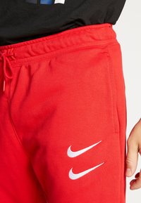 Röda Nike sweatpants har en dragsko i midjan, sidofickor och två vita logotypdetaljer på vänster lår. Mjuk bomullstextur.