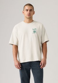 T-shirt bege de manga curta feita de algodão, com um pequeno gráfico verde da Levi's no peito e um design de ajuste descontraído.