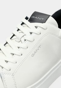 Adidași din piele albă cu o textură netedă, având șireturi albe plate, un tab de călcâi negru și logo-ul "GANT" embosat pe laterală și pe limbă.