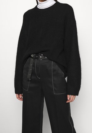 Personne portant un pull noir en tissu duveteux sur un col roulé blanc, associé à un pantalon noir taille haute avec des coutures blanches et une ceinture noire.