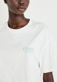 Valge puuvillane T-särk, mille vasakus rinnapiirkonnas on helesinises kirjas "CABO SAN LUCAS". Ümar kaelus ja lühikesed varrukad.
