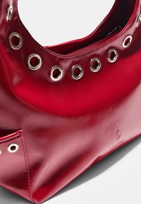 Borsa a mano in pelle rossa con occhielli rotondi in argento lungo la parte superiore. Texture liscia con chiusura a zip e una tasca laterale a contrasto.