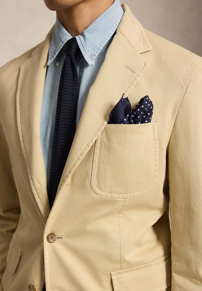Homme portant un blazer beige avec une chemise bleue, une cravate texturée foncée et un mouchoir de poche bleu marine à pois blancs.
