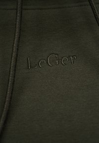 Σκούρο πράσινο ύφασμα με πλεγμένα κορδόνια και ανάγλυφο κεντημένο κείμενο "LeGer" σε νήμα ταιριαστού πράσινου χρώματος.