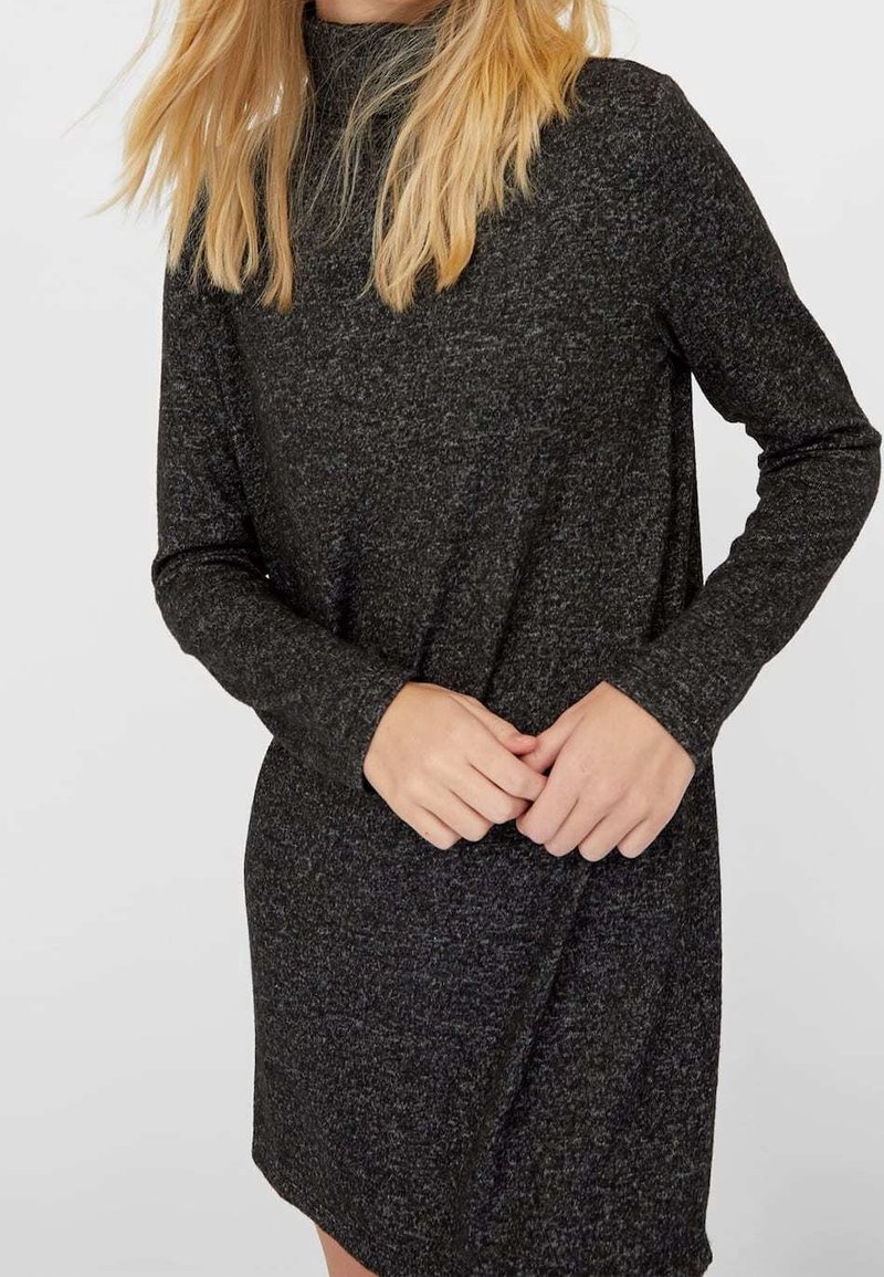 Femme portant une robe en maille à manches longues gris foncé avec un col montant, les mains légèrement jointes devant, cheveux blonds tombant sur les épaules.