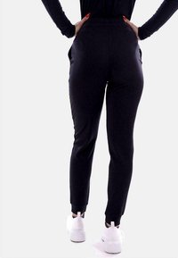 Zwarte, getailleerde joggerbroeken met zijzakken, een elastische tailleband en ribgebreide boorden. Gemaakt in combinatie met witte sneakers, waarbij de gladde stoftextuur zichtbaar is.