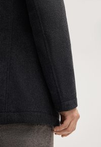 Cappotto di lana grigio scuro con una superficie texturizzata, design sagomato e manica su misura. La mano è posata vicino all'orlo, mostrando i dettagli delle cuciture.