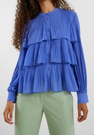 Blusa blu con pieghe e rouches a strati, chiusura con bottoni e maniche lunghe, abbinata a pantaloni verde chiaro. Tessuto morbido, leggero.