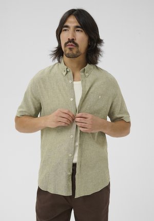 Mand med skulderlangt hår og skæg, iført en lys grøn kortærmet skjorte knapret op over en hvid tanktop, og mørkebrune bukser.