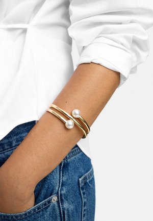 Ramię zdobi złota bransoletka z podwójnym paskiem i dwoma dużymi białymi perłami, zestawiona z podwiniętą białą koszulą i niebieskimi dżinsami.