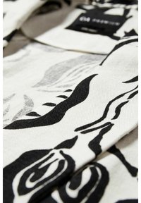 Gros plan sur un tissu blanc avec des motifs abstraits de feuilles et de lignes noires et une étiquette de vêtement noire et blanche partiellement visible.