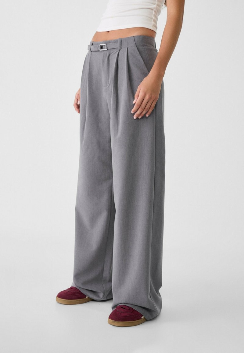 Pantalons larges gris avec des plis et une ceinture, en tissu texturé. Associés à des baskets bordeaux et un haut blanc ajusté.