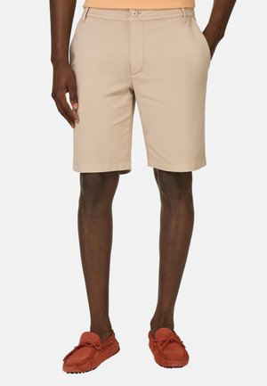 Pantaloni beige realizzati in tessuto misto cotone, con chiusura a bottone, passanti per cintura, tasche laterali e gamba dritta. Indossati con una maglietta arancione.