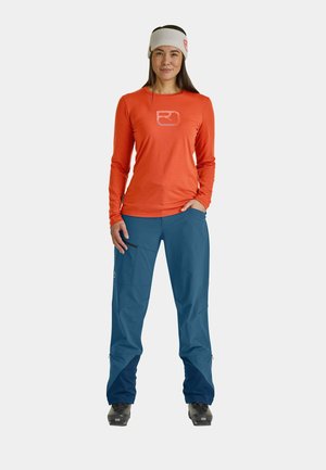 Long-sleeve orange top with logo, paired with blue pants featuring side pockets. Headband is white with textured fabric.