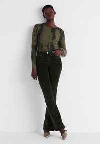 Maglietta a maniche lunghe verde oliva con motivo tie-dye, abbinata a pantaloni flare in corduroy verdi fitted. Modello con tacchi beige.