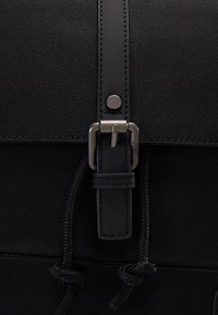 Gros plan d'un sac à dos en tissu noir avec une lanière en cuir, une boucle métallique, un bouton-pression et deux cordons noirs noués.