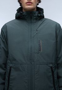 Chaqueta con capucha de color verde oscuro, con cremallera en la parte delantera, dos bolsillos laterales y detalle de logo. Textura suave con acentos de costura.