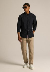 Granatowa koszula z guzikami i podwiniętymi rękawami, khaki chinosy i białe sneakersy. Model nosi ciemne okulary przeciwsłoneczne. Prosty, czysty design, bez wzorów.