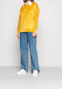 Gul manchestertröja med vit krage, tre vertikala ränder på ärmarna, kombinerad med blå vida jeans och vita sneakers.