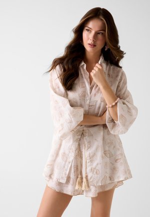 Frau mit langen welligen Haaren trägt eine hellbeige Tunika mit Muschelmuster und passende Shorts, ergänzt durch ein pinkes Armband und Ohrringe, vor weißem Hintergrund.