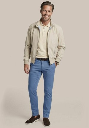Man glimlacht, draagt een beige jas en polo, lichtblauwe broek, bruine riem, horloge en donkerbruine instapschoenen, staat met één hand in de zak.