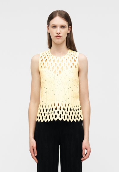 Femme portant un haut en tricot jaune sans manches avec un motif à découpes et un pantalon noir, debout devant un fond clair uni.