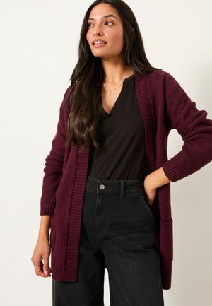Frau mit langen dunklen Haaren, die eine weinrote Strickjacke über einem schwarzen Henley-Shirt und hoch taillierte schwarze Hose trägt, steht mit einer Hand in der Tasche.