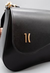 Borsa a mano in pelle nera con una texture liscia, forma curva e un logo in tono oro. Presenta una patta piegata e ferrature robuste.