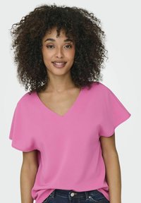 Lyserød v-hals bluse lavet af letvægtsstof med flutterærmer, vist mod en neutral baggrund, parret med mørke jeans.