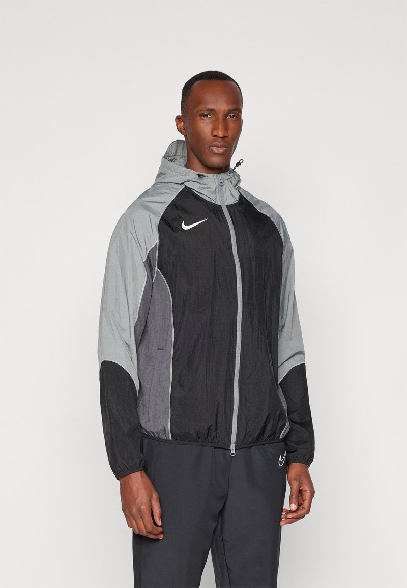 Veste légère à capuche avec un schéma de couleurs noir et gris. Comprend une fermeture éclair à l'avant, des poignets élastiques et un logo Nike sur la poitrine. Texture lisse.