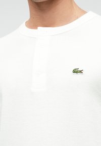 Λευκό ριγέ πουκάμισο Henley με τρία κουμπιά και μικρό κεντημένο λογότυπο κροκόδειλου σε πράσινο χρώμα στο αριστερό στήθος.