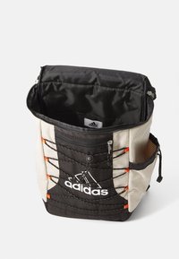 Adidas torba v bež in črni barvi z teksturirano tkanino, sprednjim žepom na zadrgo, nastavljivimi detajli iz vrvice in notranjostjo iz črnega najlona.
