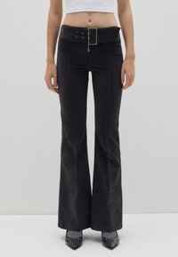 Schwarze Schlaghosen mit einem breiten Gürtel, der eine silberne Schnalle aufweist, gestickte Akzente und eine Baumwollmischung Textur. Die Schuhe sind schwarze, mit Nieten besetzte Absätze.