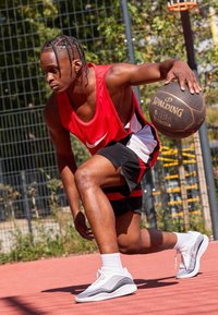 Czerwony sportowy podkoszulek z białym logo Nike, czarno-czerwone szorty, szare i białe sneakersy, trzymający piłkę do koszykówki Spalding na boisku.