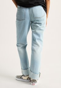 Jeans in denim blu chiaro con vestibilità rilassata, caratterizzati da orli sfilacciati e due tasche posteriori. Abbinati a scarpe slip-on a scacchi bianche e nere.