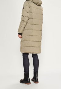 Person wearing a long beige quilted coat with hood, dark skinny jeans, and black ankle boots, standing against a plain white background.